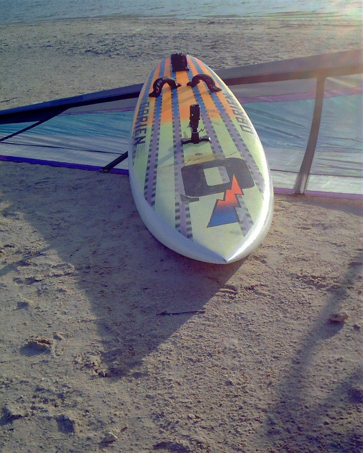 Mike Kimbro's Aerotech 7.5 sail behind his vintage 1989 O'Brien Epoxy Lite 9.9 windsurfer, as the sun drops low at Corpus Christi's Bird Island Basin sailing and fishing area.
